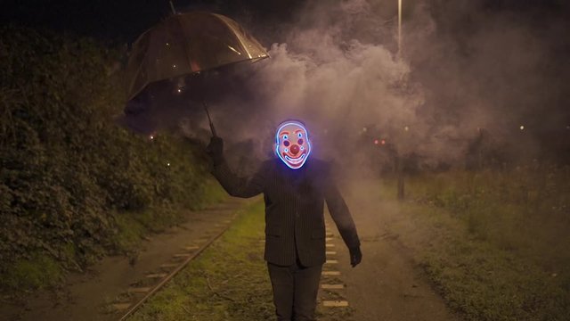 Man with clown mask and smoking umbrella is walking over a railroad at night.