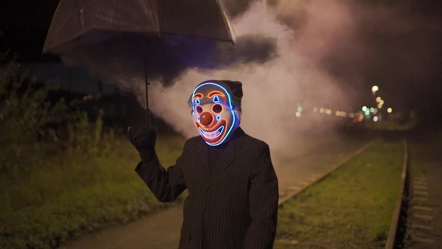 Man with clown mask and smoking umbrella is walking over a railroad at night.