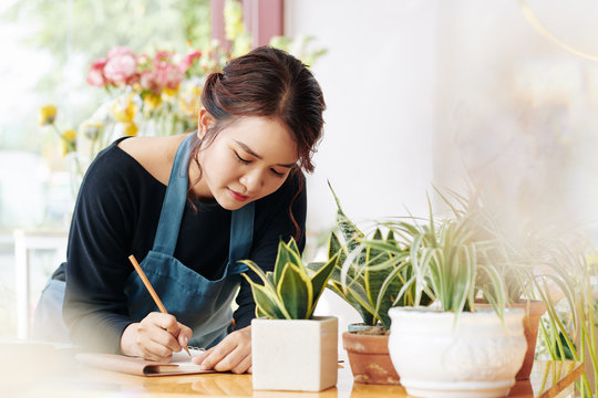 Young Asian flower shop owner writing list of plants and pots she needs to order