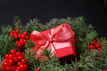 New Year Christmas Xmas 2020 holiday celebration red present gift box with satin pink bow, immersed in the needles of a Christmas tree decorated with red berries. Dark festive composition.