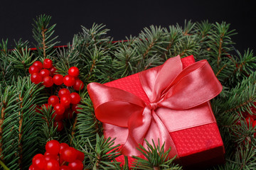 New Year Christmas Xmas 2020 holiday celebration red present gift box with satin pink bow, immersed in the needles of a Christmas tree decorated with red berries. Dark festive composition.