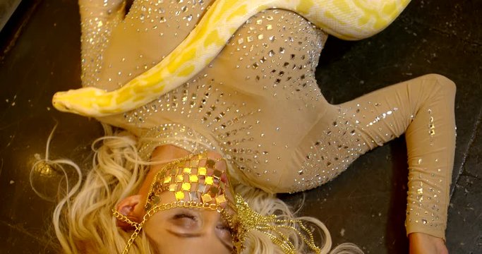 the blonde is lying on the floor and stroking the snake. the girl is dressed in a gold jumpsuit with rhinestones. a woman's face is half hidden by a gold ornament. the view from the top