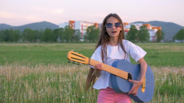 leisure activity happy hippie retro little girl in sunglasses playing denim acoustic guitar on summer outdoor green field
