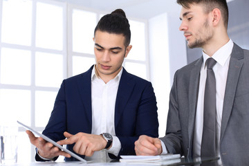 Fototapeta premium Two businessmen or partners working with tablet computer at the desk in modern office. Headshot of arab male entrepreneur or manager with colleague at workplace. Multi ethnic teamwork, partnership and