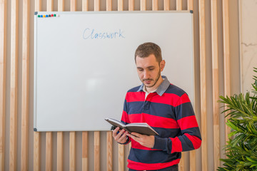 Young man standing at the blackboard and looking at a notebook