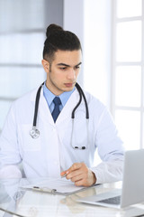 Arab doctor man using laptop computer while filling up medication history records form at the glass desk in medical office or clinic. Medicine and healthcare concept