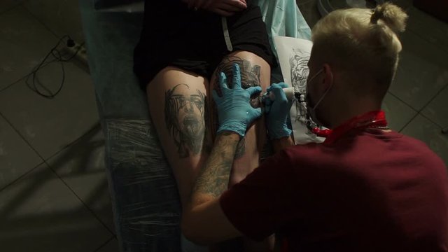 Close-up of a tattoo artist doing a tattoo on a young woman's leg in the salon. Professional tattooist doing tattooing in studio. Skin art close-up. Slow motion.
