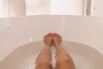 Woman taking a hot bath selfie in tub home cozy relaxation in winter night. POV of feet and legs in...