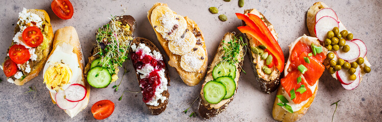 Open toasts with different toppings on gray-brown background, top view.  Flat lay of crostini with banana, pate, avocado, salmon, egg, cheese and berries.