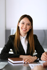 Happy smiling business woman headshot. Unknown businesswoman sitting at meeting or negotiation in modern office with colleagues at the background. Teamwork and partnership concept
