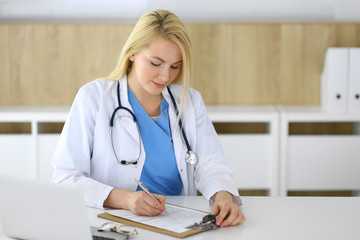 Doctor woman at work while sitting at the desk in hospital or clinic. Blonde cheerful physician filling up medical records form. Data and best service in medicine and healthcare