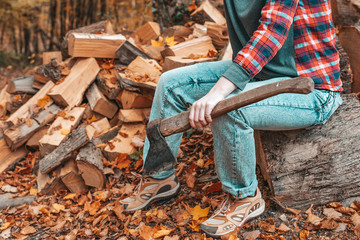 Preparation for the heating season. A young woman with an axe in her hands sits on a log. In the background, a pile of firewood. Close up