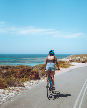 Woman Riding A Bicycle Along A Bike Path With The Beautiful Blue Ocean As The Backdrop, At Rottnest Island In Perth, Western Australia.