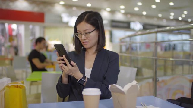 Waist-up shot of cheerful young Chinese woman in blazer and glasses sitting at table in food court of mall, talking on video call and sharing pictures of shopping bags, fast food and her happy face