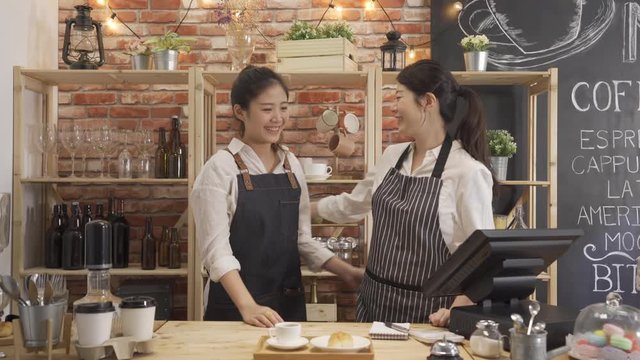 two young asian women coworker working at cafe bar face camera smiling attractive. girls barista standing in counter close hugging each other joyful laughing look at lens in modern coffee shop.