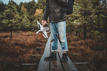 Young hipster in blue denim jeans is standing on wooden path with drone in hand.