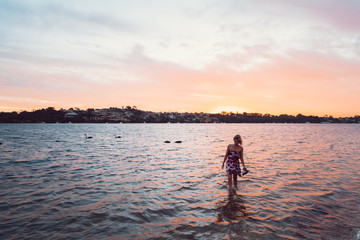 Beautiful colourful sunset at Point Walter, woman in summer dress walking through the water on the sand bank with swans and Perth city in the background.