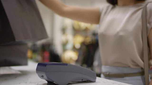 Lockdown arc shot of unrecognizable woman touching smartphone onto payment terminal at checkout counter of fashion store, taking shopping bags and leaving