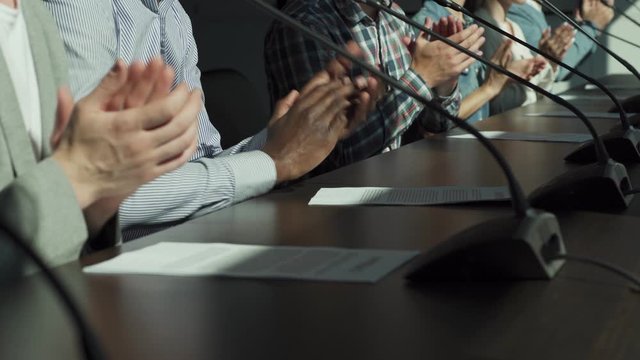 Closeup hands of multiethnic unrecognizable business people applauding to speaker sitting at desk in a row with contract papers and microphones in front of them during business conference