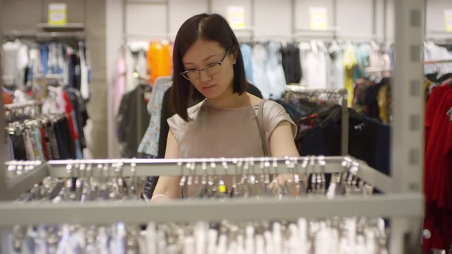 Medium tracking shot of young Asian woman walking around clothing shop, looking through garments on rails, then picking up skirt on hanger and checking it out