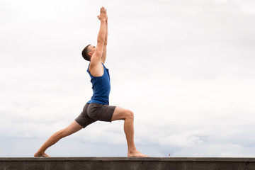 attractive young man doing yoga warrior pose on the beach on sky background