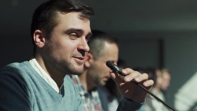 Tracking right closeup of smiling young man speaking into microphone in business meeting. Businessman reading his speech, side view