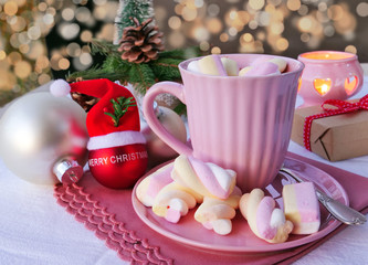Hot chocolate with marshmallows in pink mug on Christmas table with candle on bokeh background.