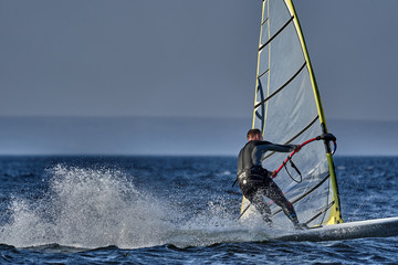 A male athlete is interested in windsurfing. He moves on a Sailboard on a large lake on an autumn day.