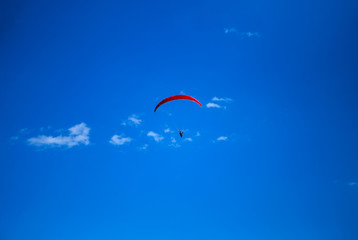 The red paraglider flies high above, with only blue skies and a few small clouds behind.