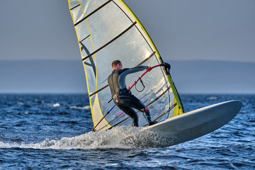 A male athlete is interested in windsurfing. He moves on a Sailboard on a large lake on an autumn day.