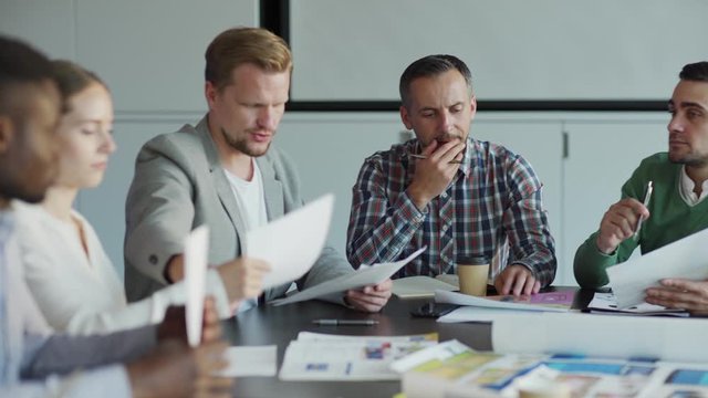 Tracking medium shot of business team sitting at table in meeting and discussing project. Man giving papers with data to colleagues