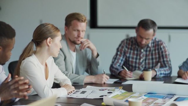 Tracking shot of diverse business team discussing market analysis sitting at desk in meeting. Young woman holding document with charts and explaining statistics to colleagues