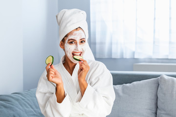 Beautiful young woman with facial mask on her face holding slices of fresh cucumber. Skin care and treatment, spa, natural beauty and cosmetology concept