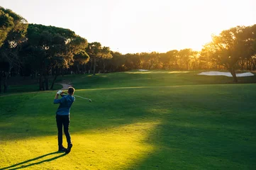 Rolgordijnen Male golf player teeing off golf ball from tee box to beautiful sunset © karrastock