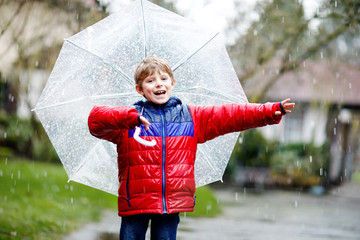 Beautiful little kid boy on way to school walking during sleet, rain and snow with an umbrella on cold day. Happy and joyful child in colorful fashion casual clothes.