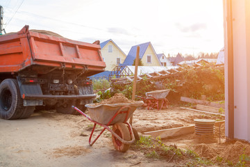 Obraz premium wheelbarrow with sand and shovel in foreground and dump truck preparing to dump sand on ground in background