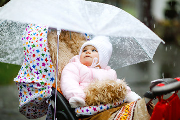 Cute little beautiful baby girl sitting in the pram or stroller on cold day with sleet, rain and snow. Happy smiling child in warm clothes, fashion stylish baby coat. Baby with big umbrella