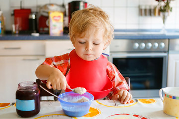 Adorable toddler boy eating healthy porrige from spoon for breakfast with berry jam. Cute happy...