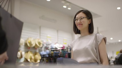 Waist-up shot of smiling Asian woman scanning her palm on payment terminal at checkout counter of clothing store, taking shopping bags from sales assistant, thanking, saying goodbye and leaving