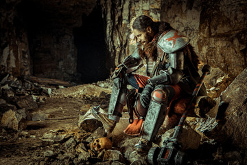 Powerful knight in the armor with the hammer is sitting on the stone. Dungeon on the background.