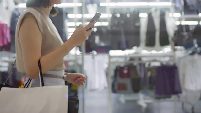 Tracking shot of unrecognizable woman with short hair walking through shopping mall past glass window displays, using smartphone and carrying many paper bags with purchases