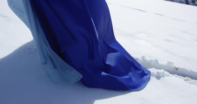 Woman in blue dress is walking through snowland. Footprints on the snow and hem clouseup.