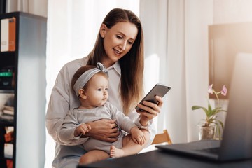Little girl sitting on mother's knees and looking at screen of mom's smartphone. Young woman and daughter making video call in messenger app on mobile phone. Girl texting messages and comments in blog