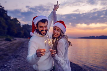 Cheerful caucasian happy couple in love holding sparklers and dancing on shore near river. Both are dressed in white sweaters and having santa hats. Selective focus on sparklers.