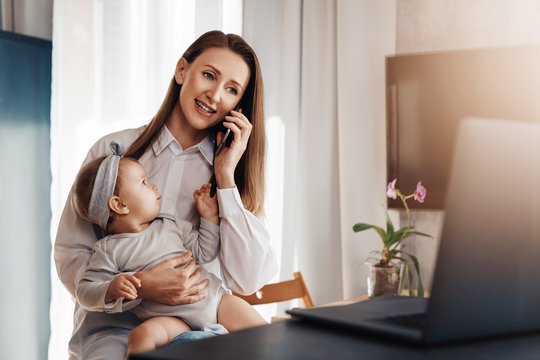 Young woman making phone call on her mobile, baby daughter sitting on knees and watching at mom. Mother working on laptop remotely while taking care of child at home. Freelance career for young moms