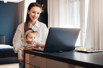 Mother with little daughter sitting at home by window and watching funny cartoons for children on laptop. Home-based freelance work for young parent, vacancies for experts and specialists with kids