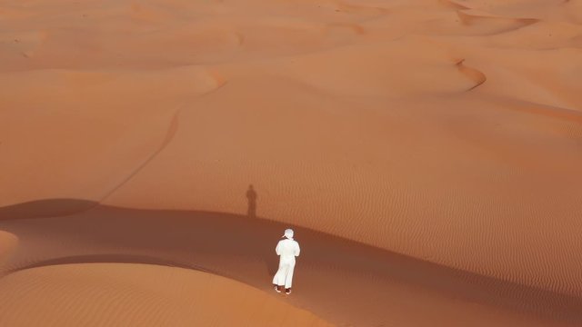 Rising drone shot of man in traditional arab clothing (thawb) stands on a dune in a desert. 