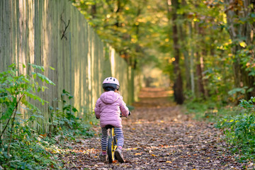 Young girl in warm clothing on balance bike riding along an autumnal footpath besides a high wooden fence in the forest. Seen in October in Germany, Bavaria