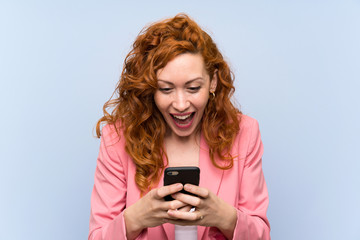 Redhead woman in suit over isolated blue wall surprised and sending a message
