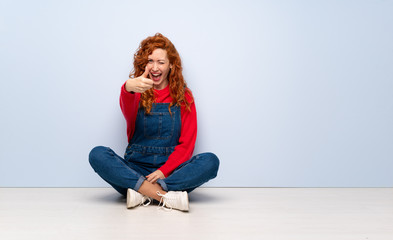 Redhead woman with overalls sitting on the floor with thumbs up because something good has happened